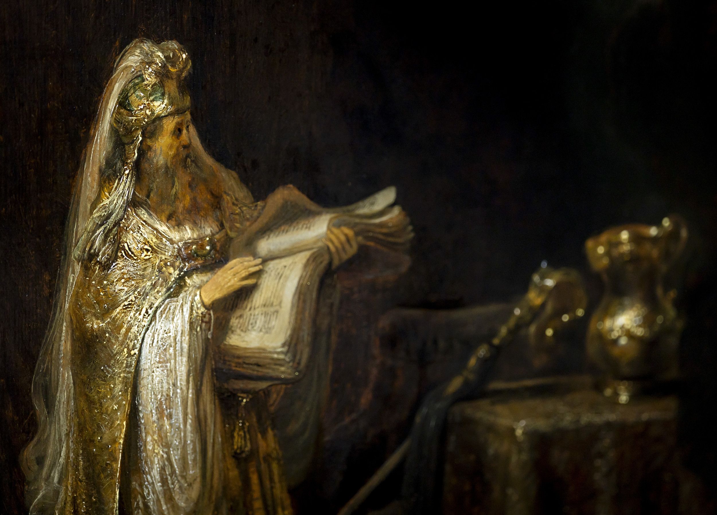 Foto van een schilderij van een oude man in goudkleurig oosters gewaad en met een opengeslagen boek in zijn hand. 