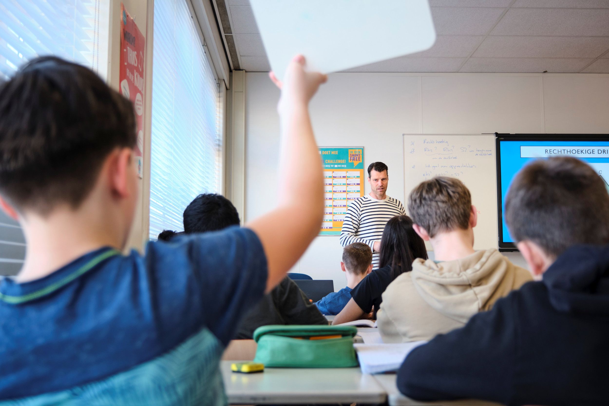 Een docent met een zwart-wit gestreept shirt staat voor de klas. Leerlingen zijn op de rug gefotografeerd. Een van de leerlingen steekt een zogeheten wisbordje de lucht in.