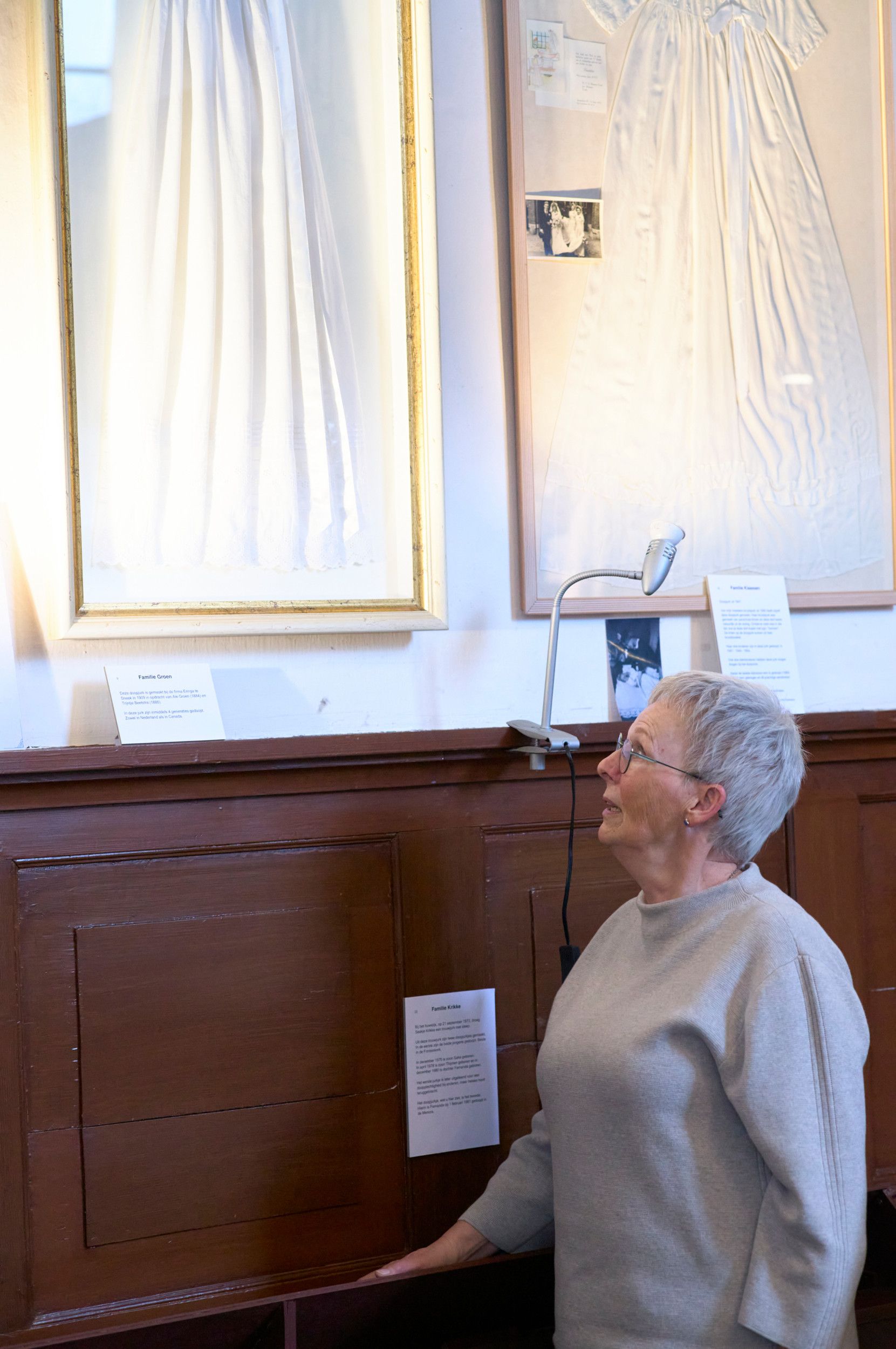 Een oudere vrouw met kort grijs haar, bril, kijkt naar de doopjurk uit haar familie.