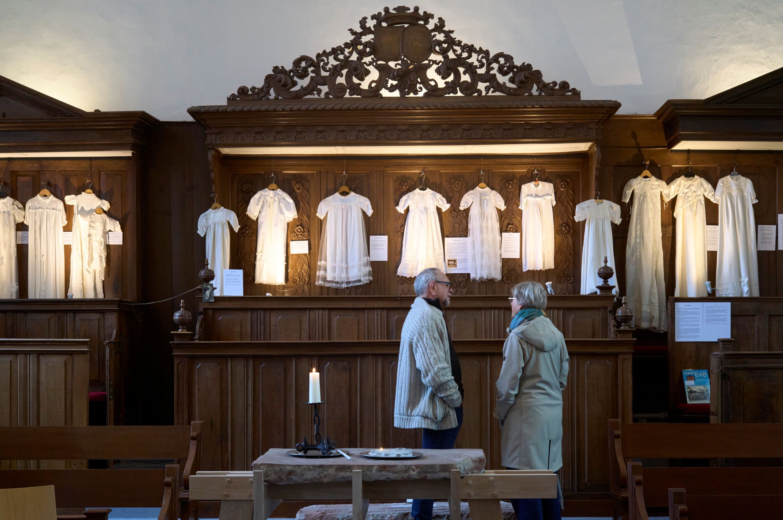 Een oudere man en vrouw in gesprek bij een aantal doopjurken.