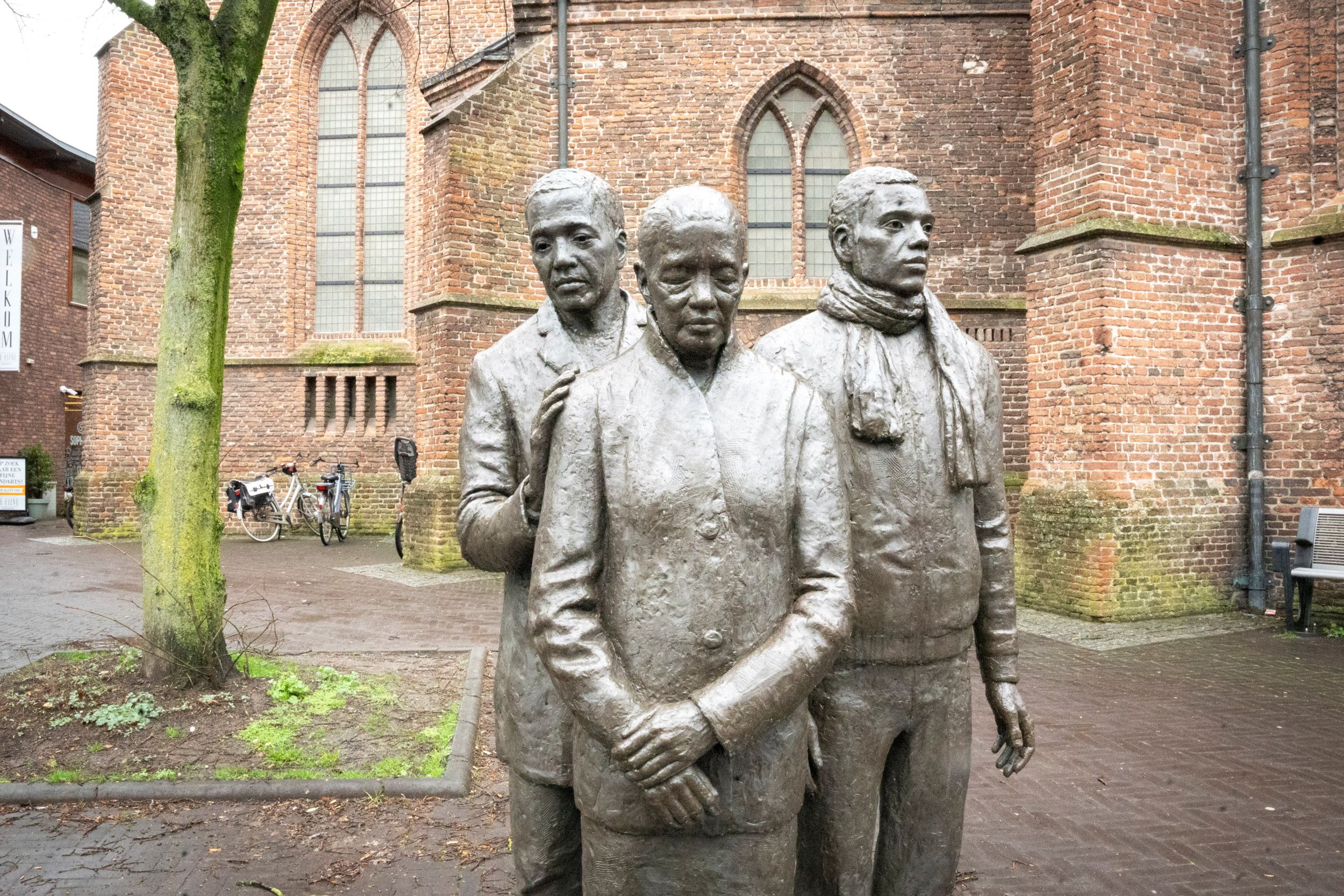 Drie bronzen figuren voor een oude kerkmuur. De zoon legt de hand op de schouder van zijn moeder.