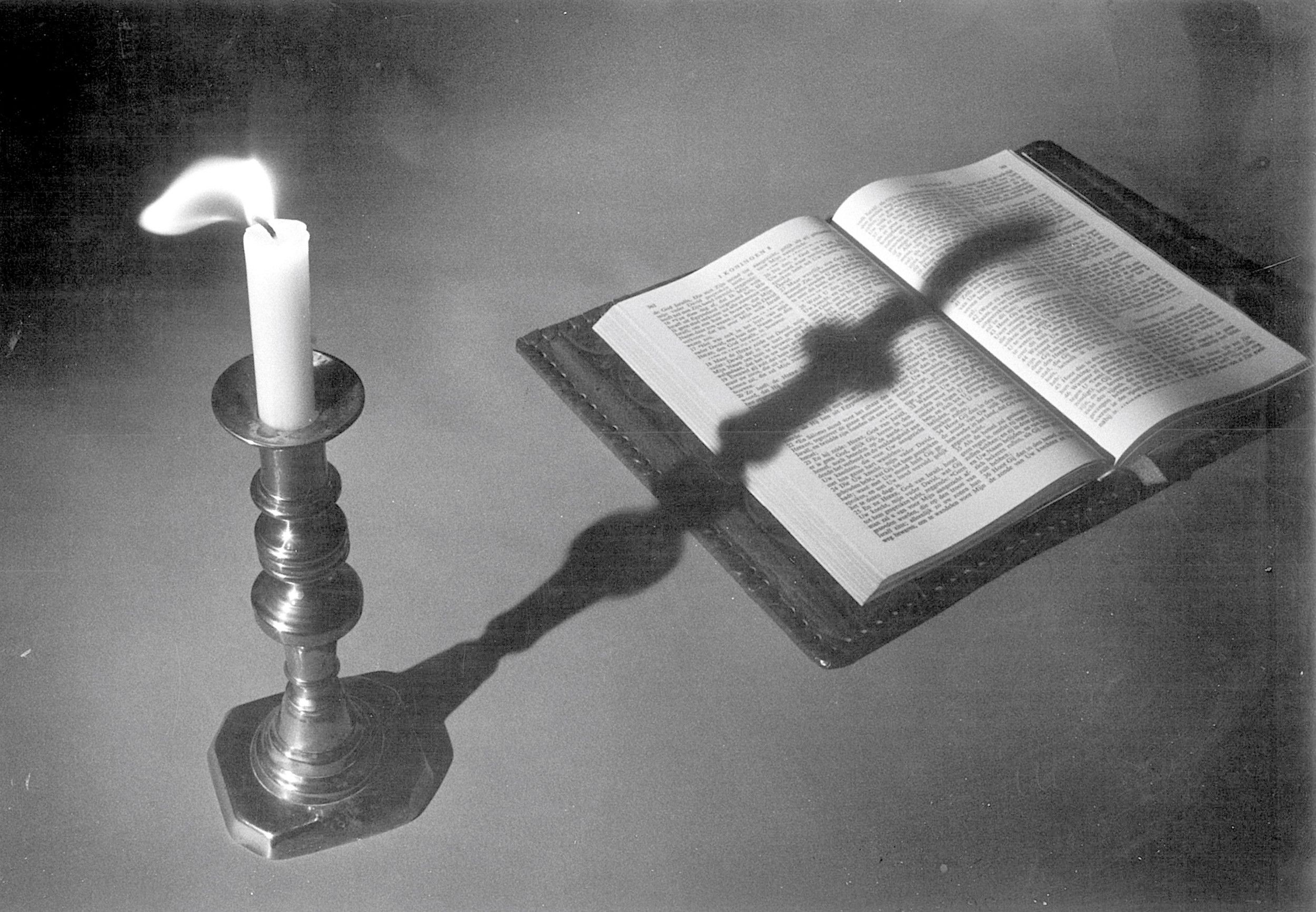 Een zwart-witfoto van een kaars die zijn schaduw werpt op een opengeslagen Bijbel.
