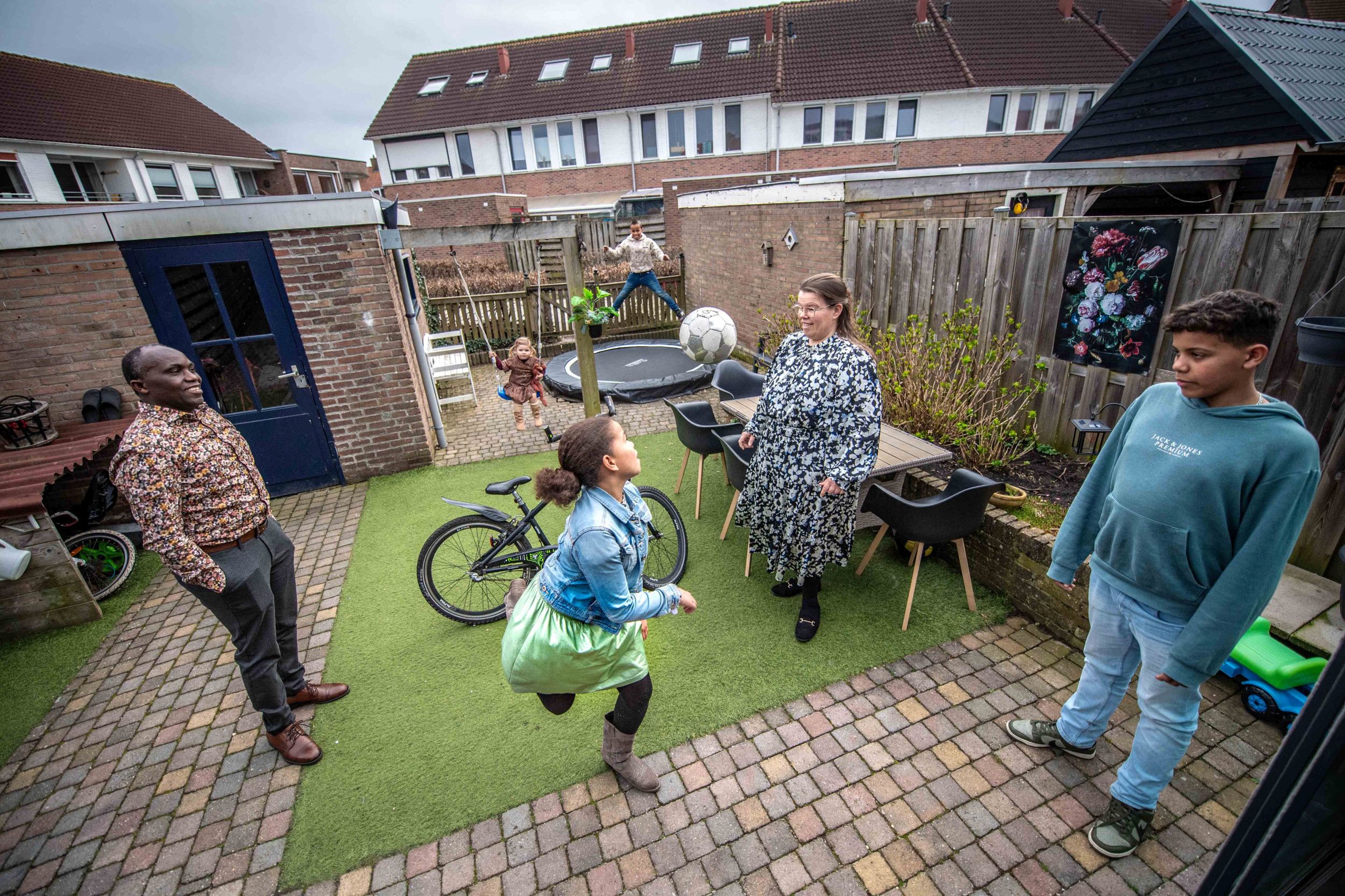 Betegelde achtertuin met trampoline en schommel. Kinderen zijn aan het spelen met een bal, ouders staan erbij.