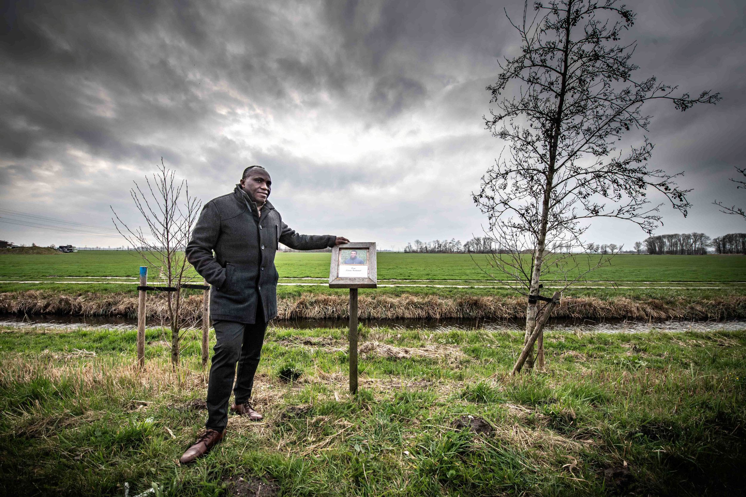 Man met donkere huidskleur en in donkere kleren bij het bordje dat herinnert aan het ongeluk en overlijden van zijn zoon.