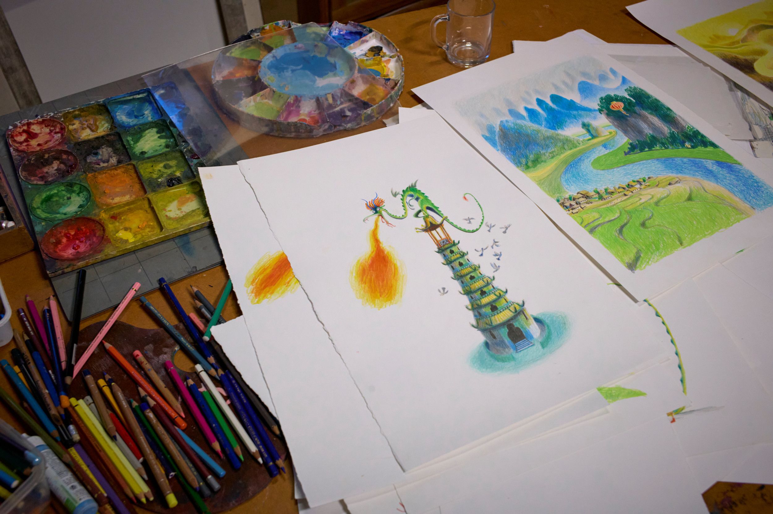 Twee kleurrijke getekende illustraties van een landschap en een toren met een draak erop. Links ervan een hoopje potloden en twee verfpaletten.