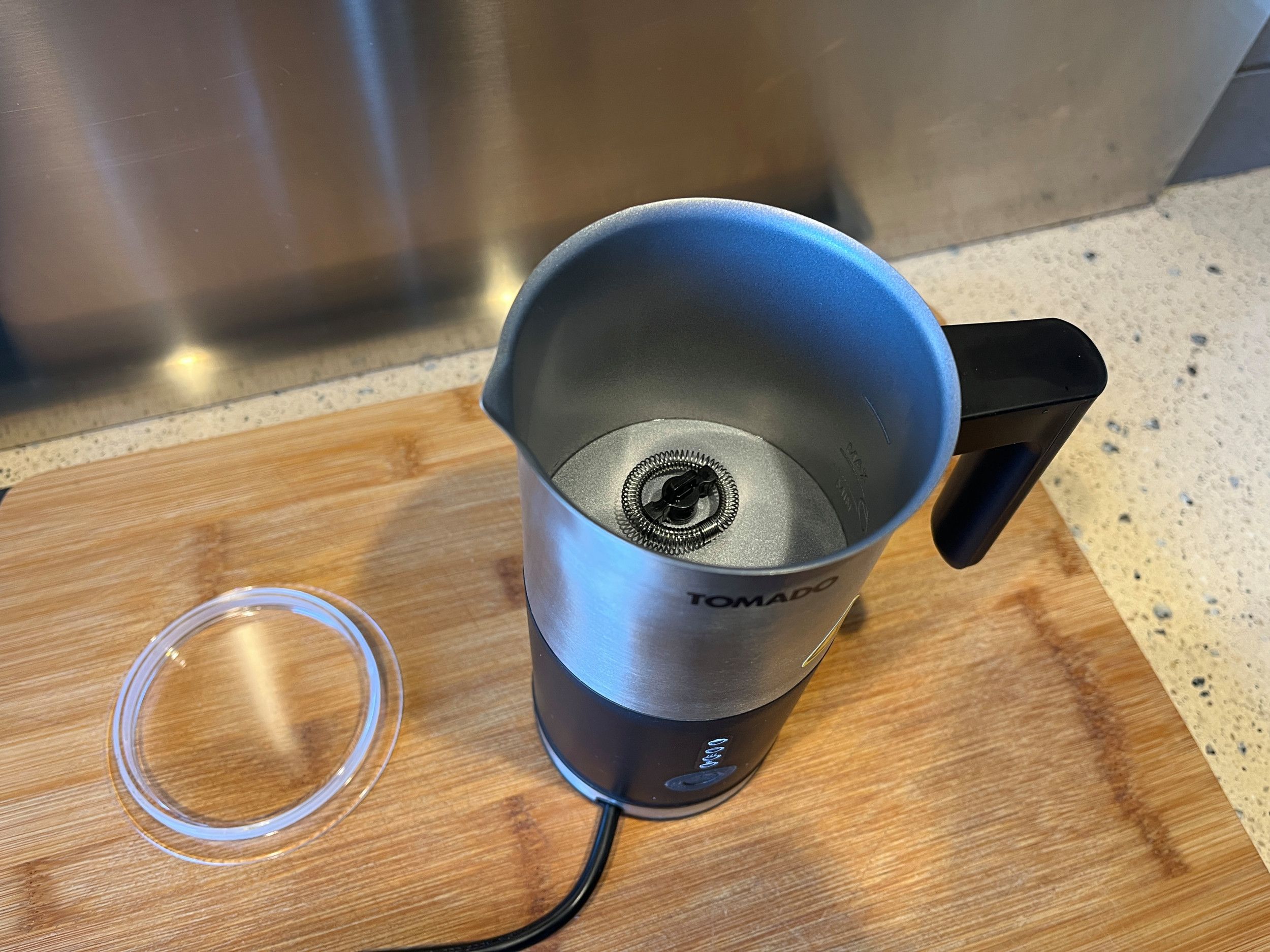 op een houten snijplank staat een melkopschuimer, een kannetje dat bovenaan van staal is, en onderaan van zwart plastic. In het kannetje zit een ijzeren spiraatje bevestigd. naast het kannetje ligt een transparant dekseltje