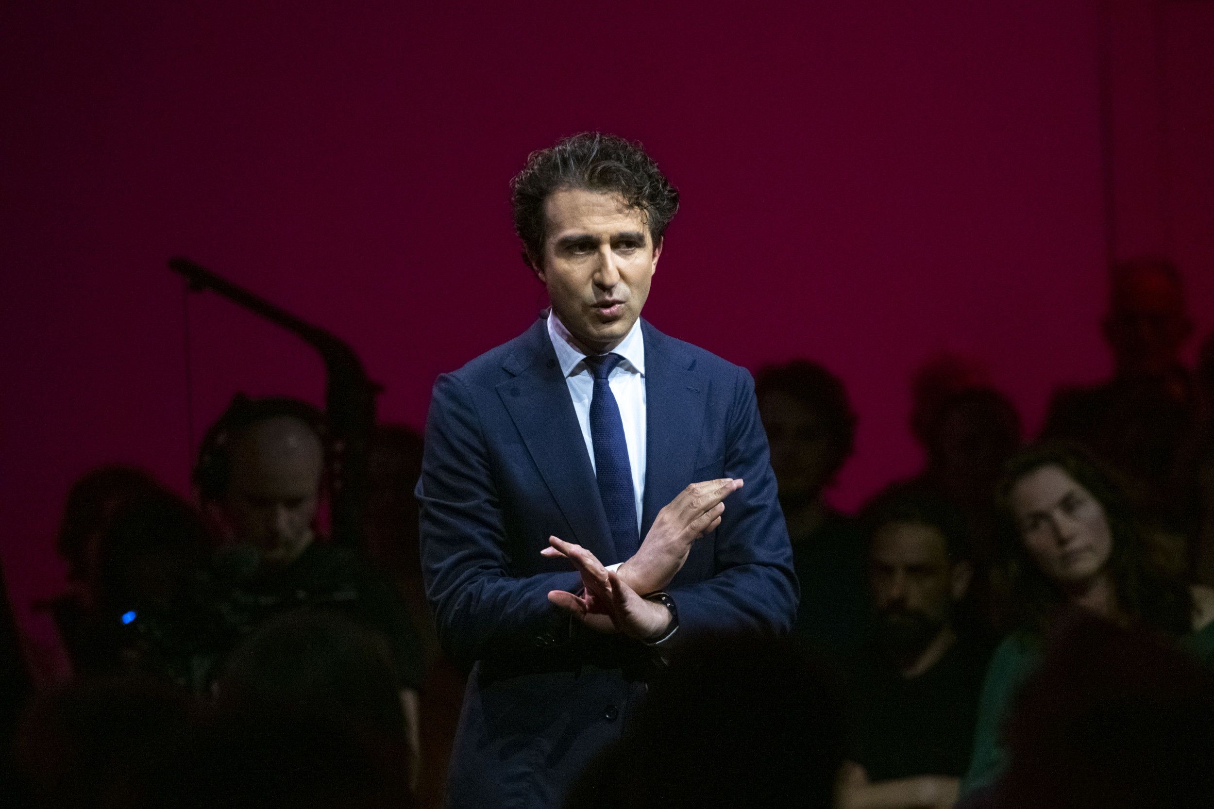 Man met donker haar en donkerblauw pak die zijn handen gekruist voor zijn lichaam houdt.