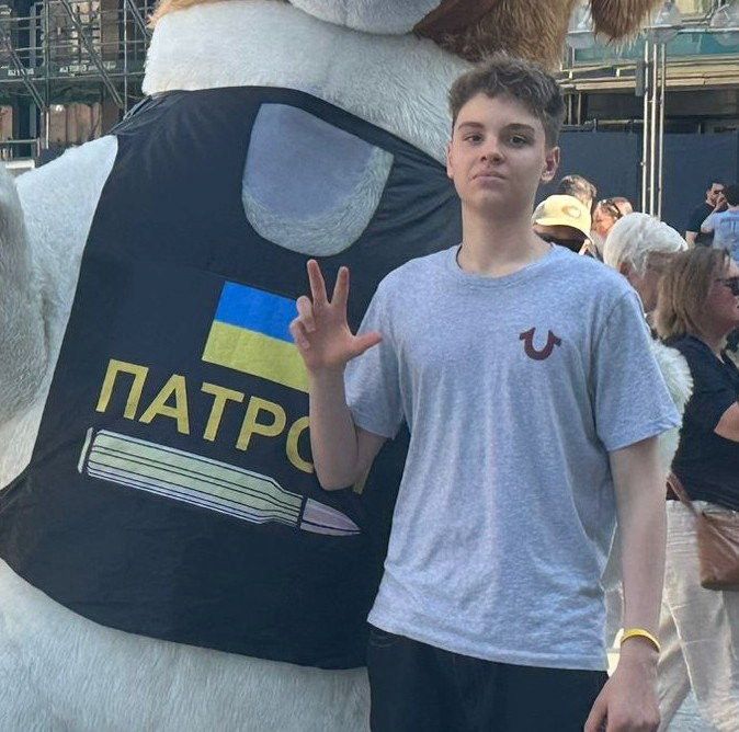 Een jongen in een grijs shirt en met bruin haar steekt drie vingers op. Naast hem is een Oekraïense vlag zichtbaar.