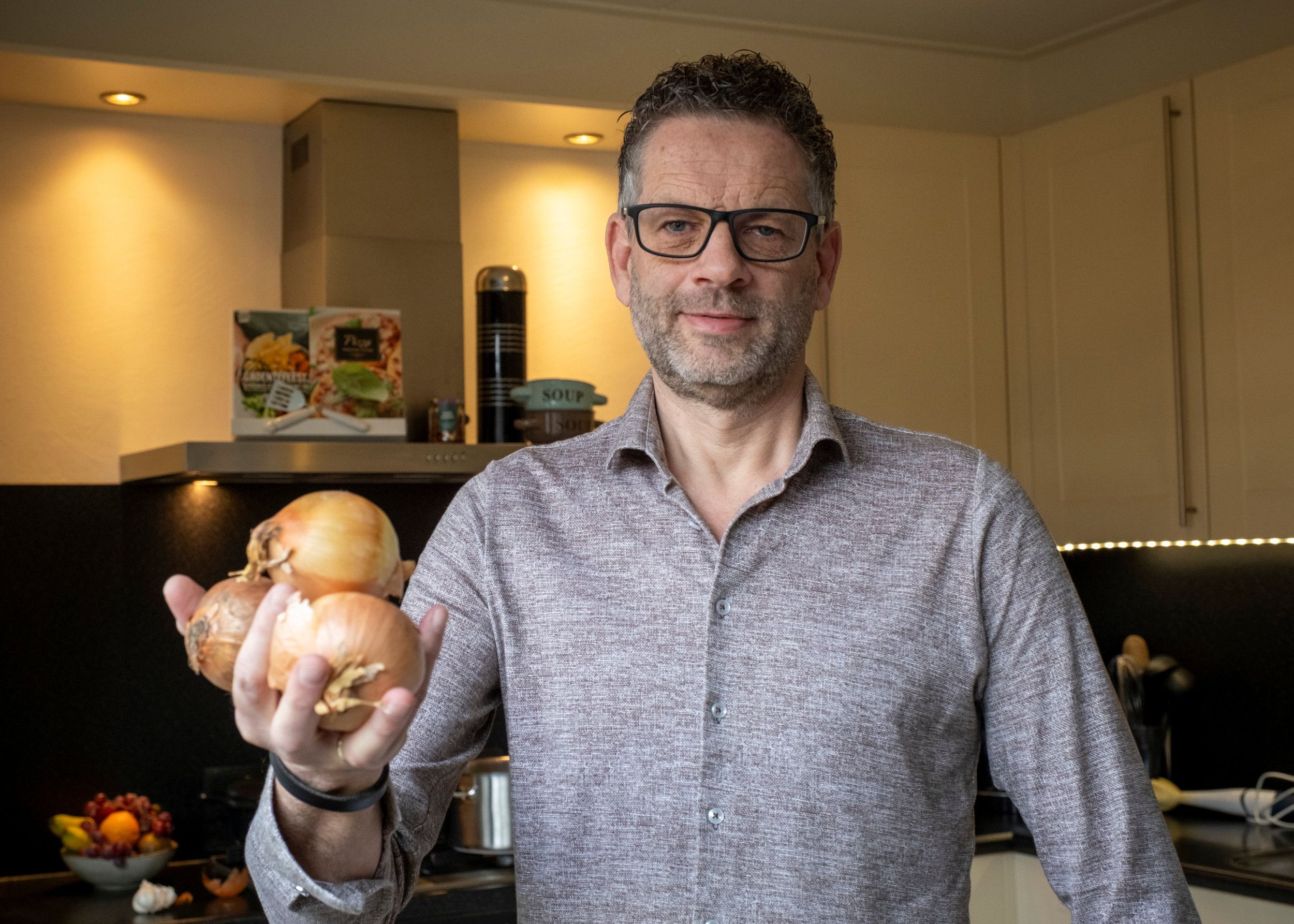 Een man van middelbare leeftijd staat in een keuken. In zijn hand houdt hij een paar uien vast. Op de achtergrond is een afzuigkap te zien. Op de rand daarvan wat soepkommen, een thermosfles en een paar kookboeken.