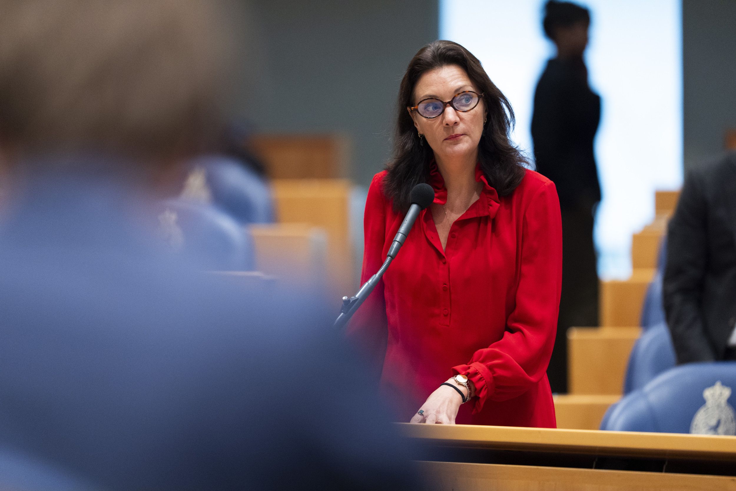 Mevrouw met donker haar, een bril en een rode jurk staat achter een microfoon in de zaal van de Tweede Kamer.