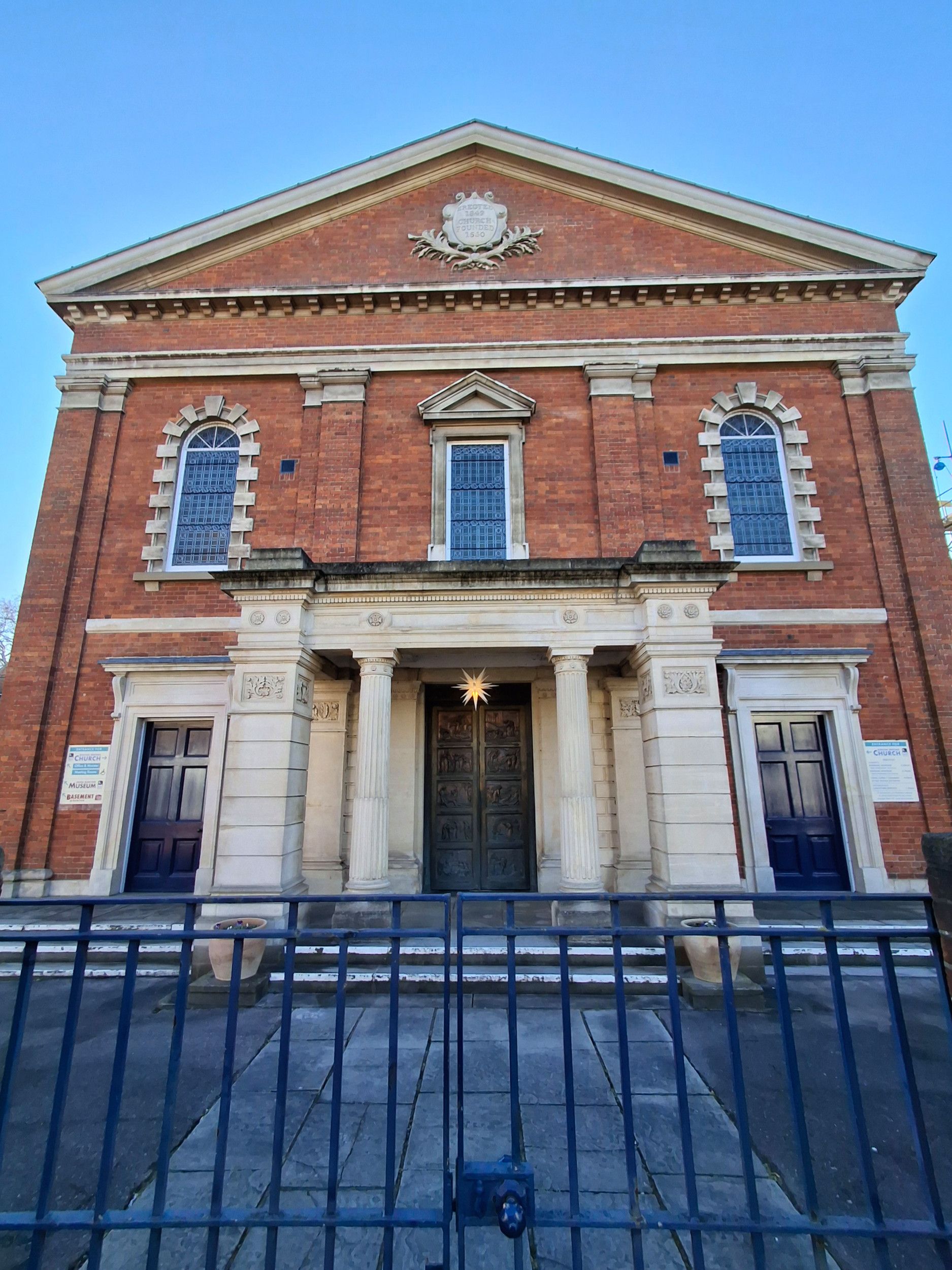 Foto van een oud kerkgebouw, met een entree die steunt op zuilen. Ervoor een plein en een hekwerk.