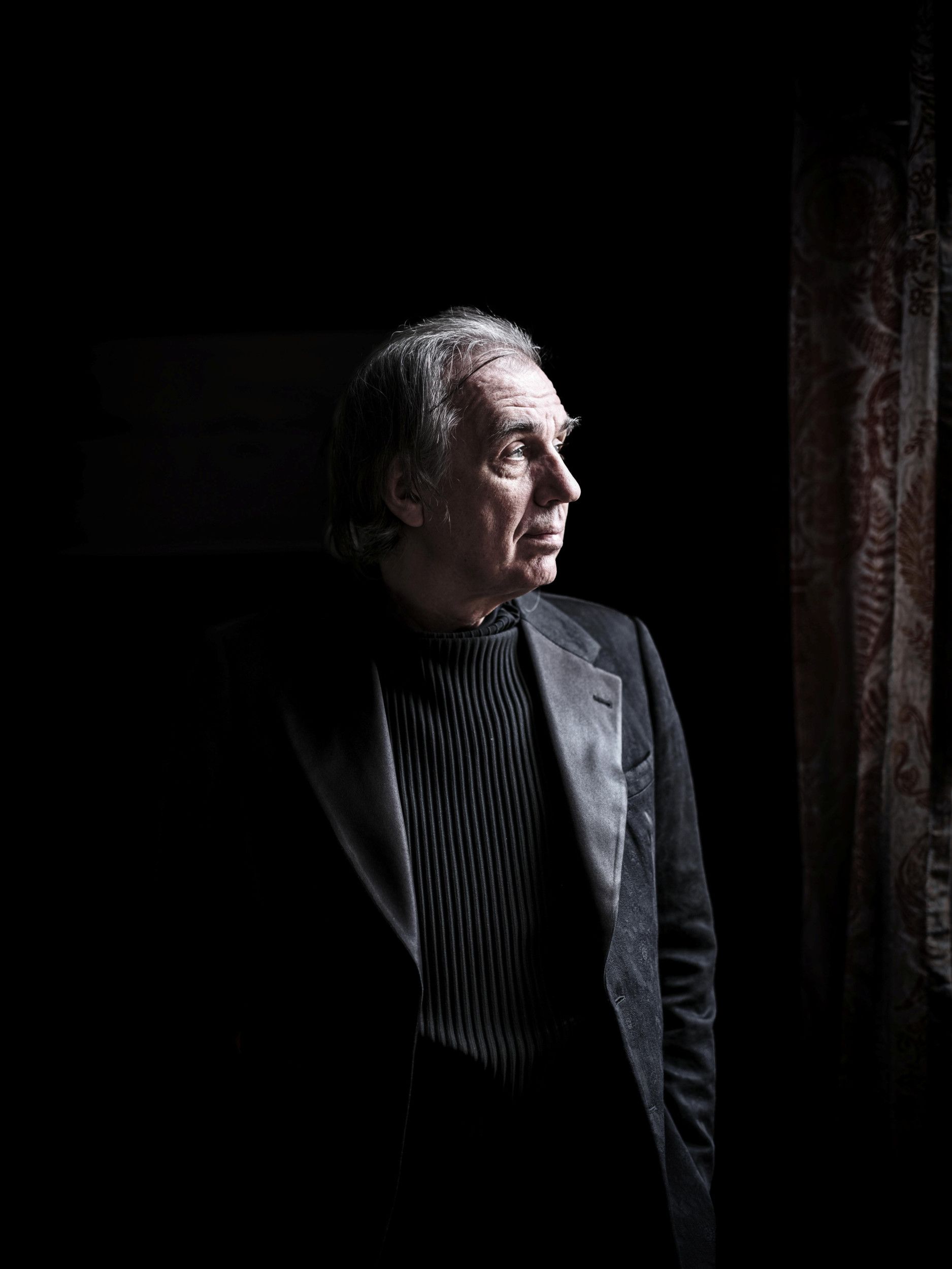 Oudere man met grijs haar en zwarte kleding tegen een zwarte achtergrond. Daglicht valt door een raam op zijn gezicht.