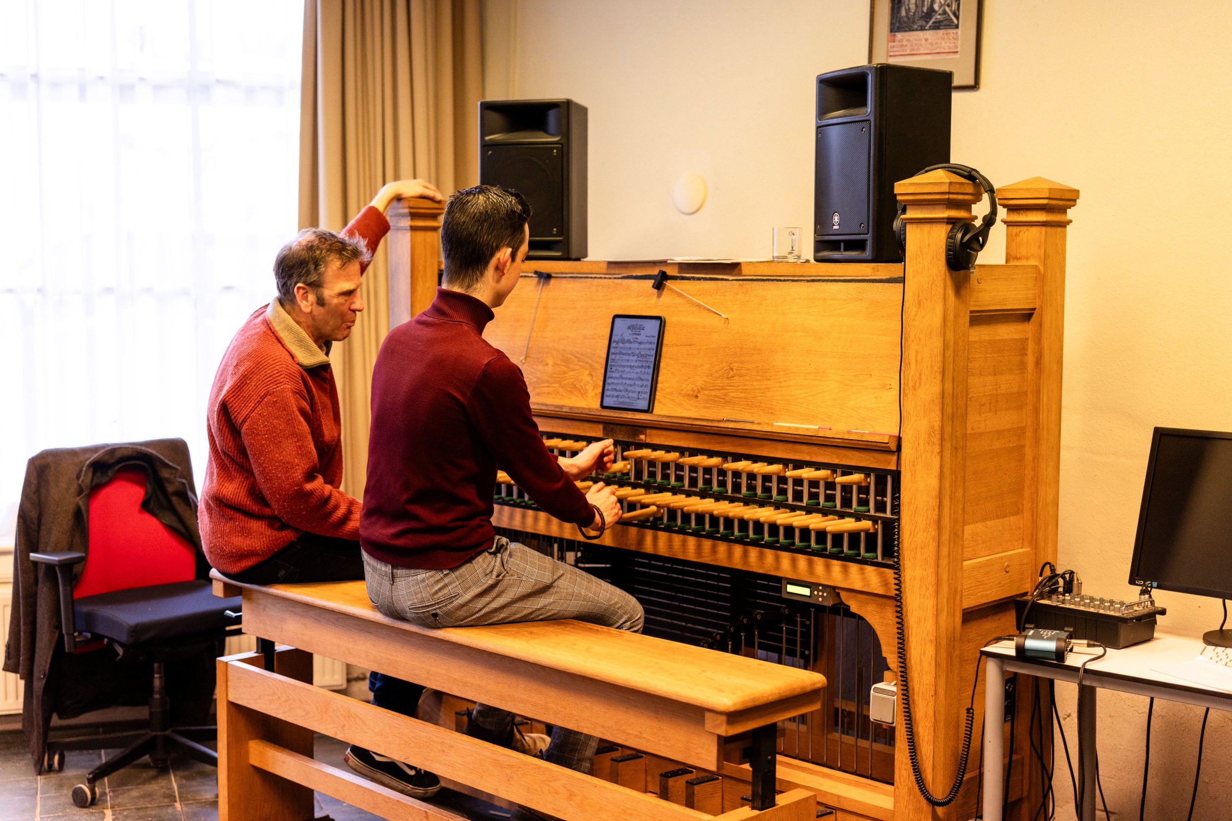 Een jongeman speelt op een oefenbeiaard in een kamer. Een man, zijn docent, zit links van hem op de bank van de oefenbeiaard en kijkt aandachtig toe.