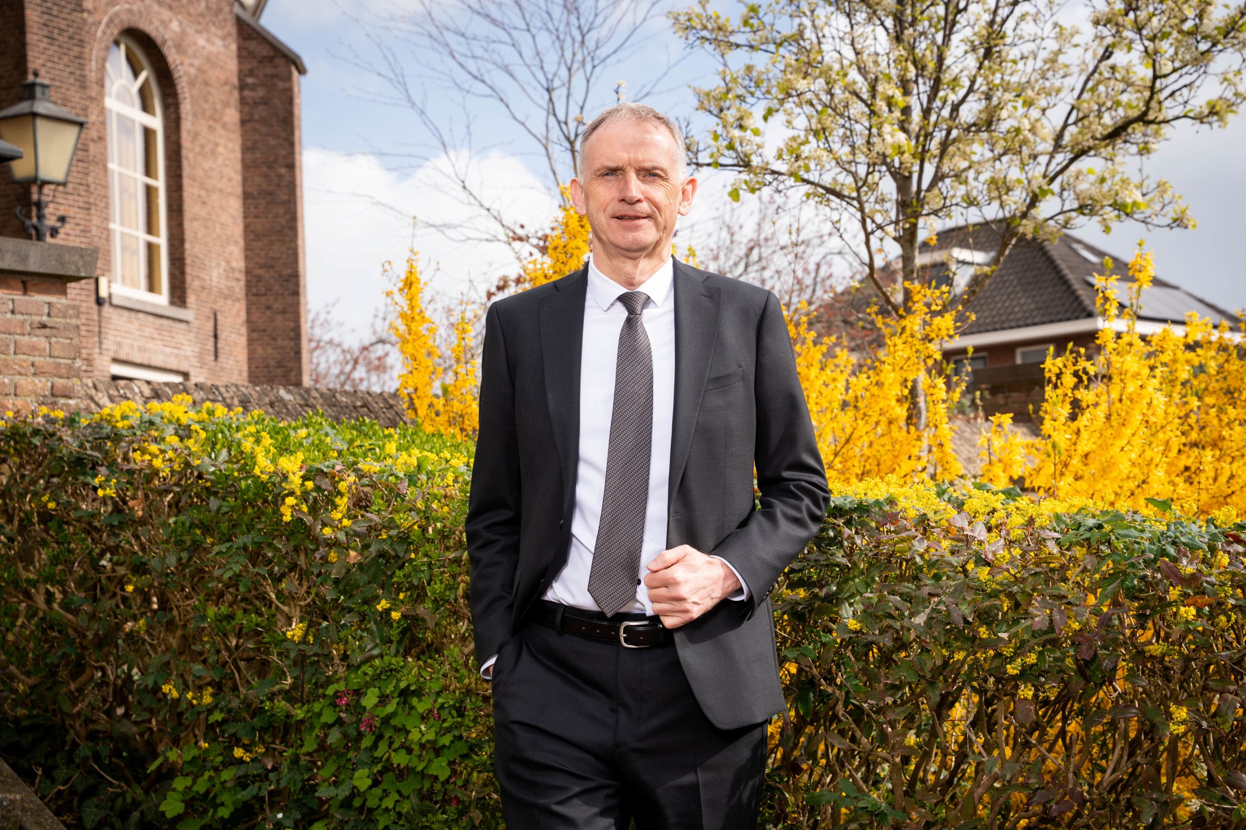 Man in een zwart pak met wit overhemd en stropdas staat buiten voor een heg.