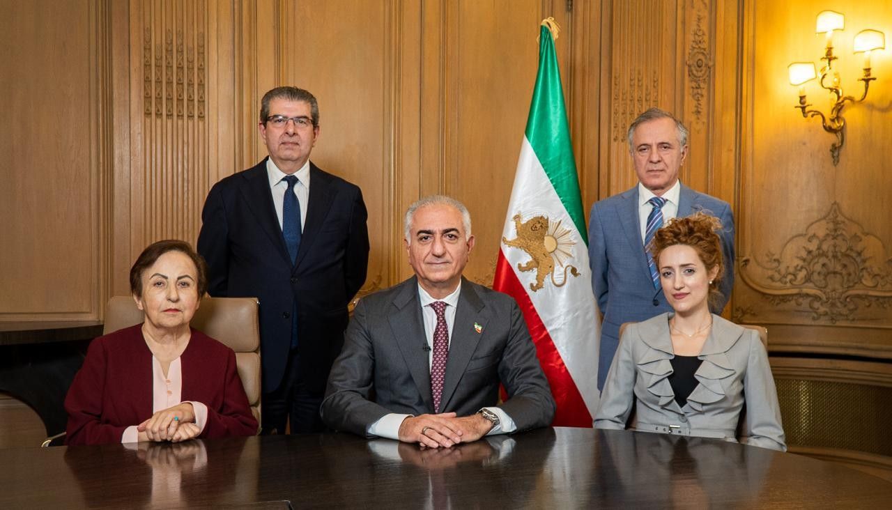 Twee vrouwen en drie mannen achter een tafel. Bij hen een Perzische vlag.  