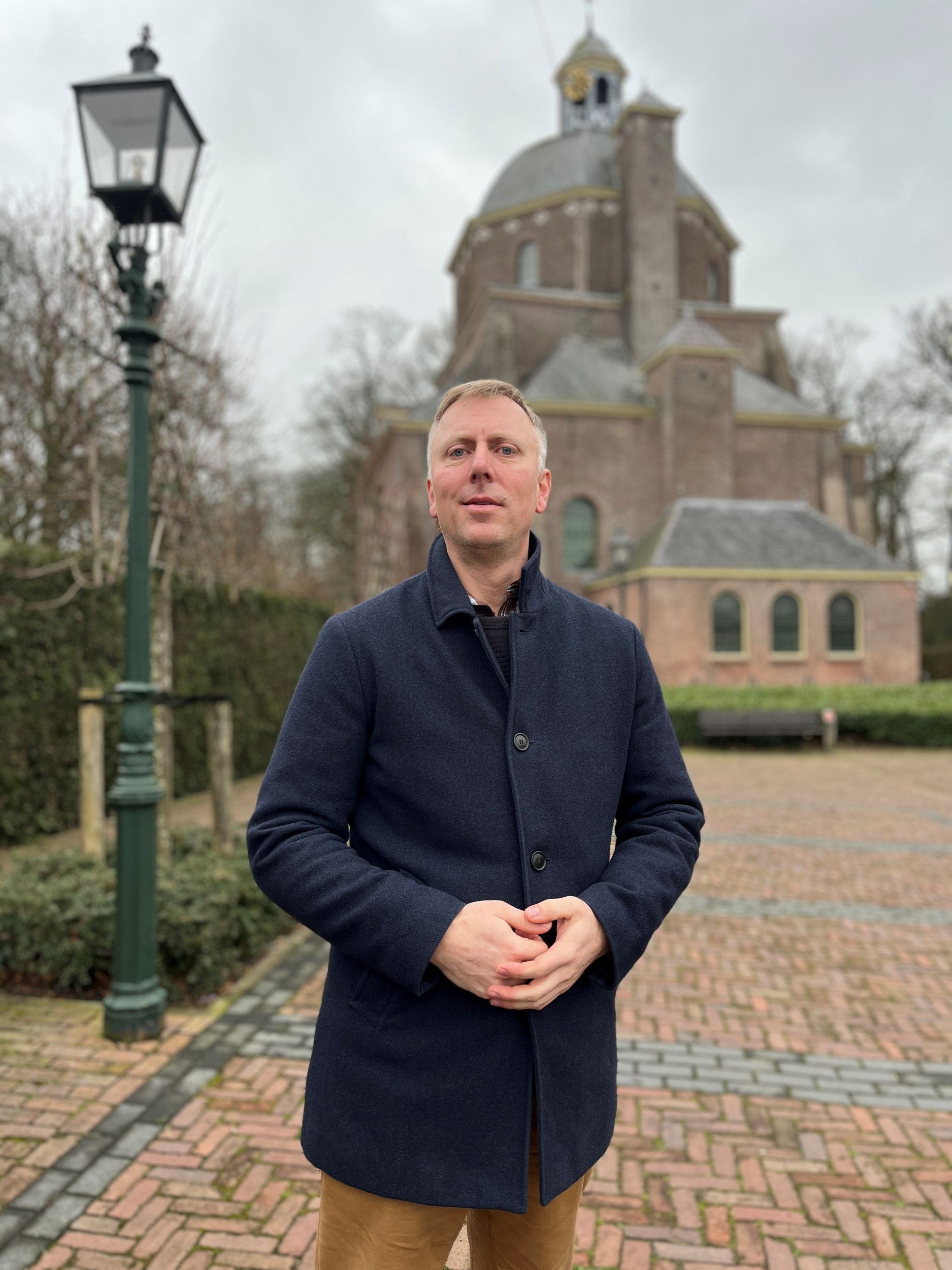 Man met ovaal gezicht, blond, grijzend haar, blauwe ogen, donkerblauwe wollen jas aan, staat voor een kerkgebouw.