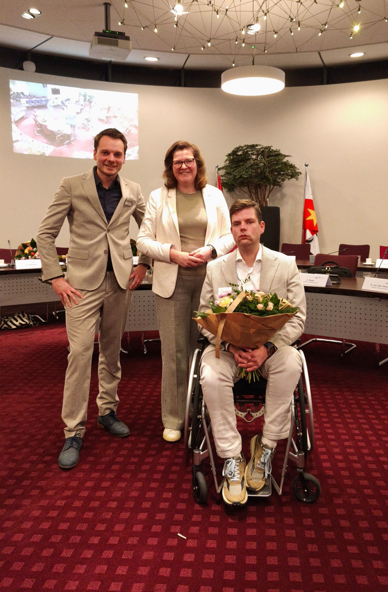 Drie mensen in nette kleding, een van de mannen zit in een rolstoel. Hij heeft een bos bloemen op schoot.