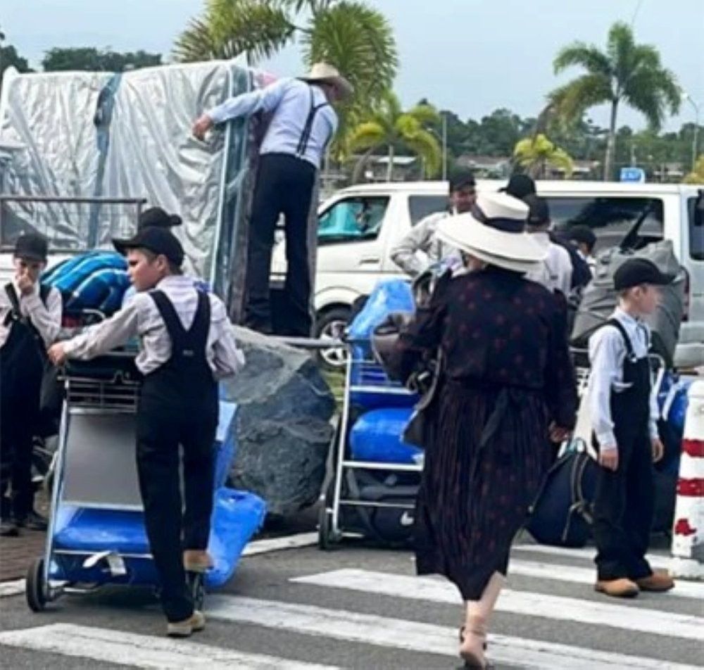 Jonge jongens in een soort donkere tuinbroeken, witte blouse, petje op, en hun ouders. Ze zijn bezig spullen uit hun busjes te laden.