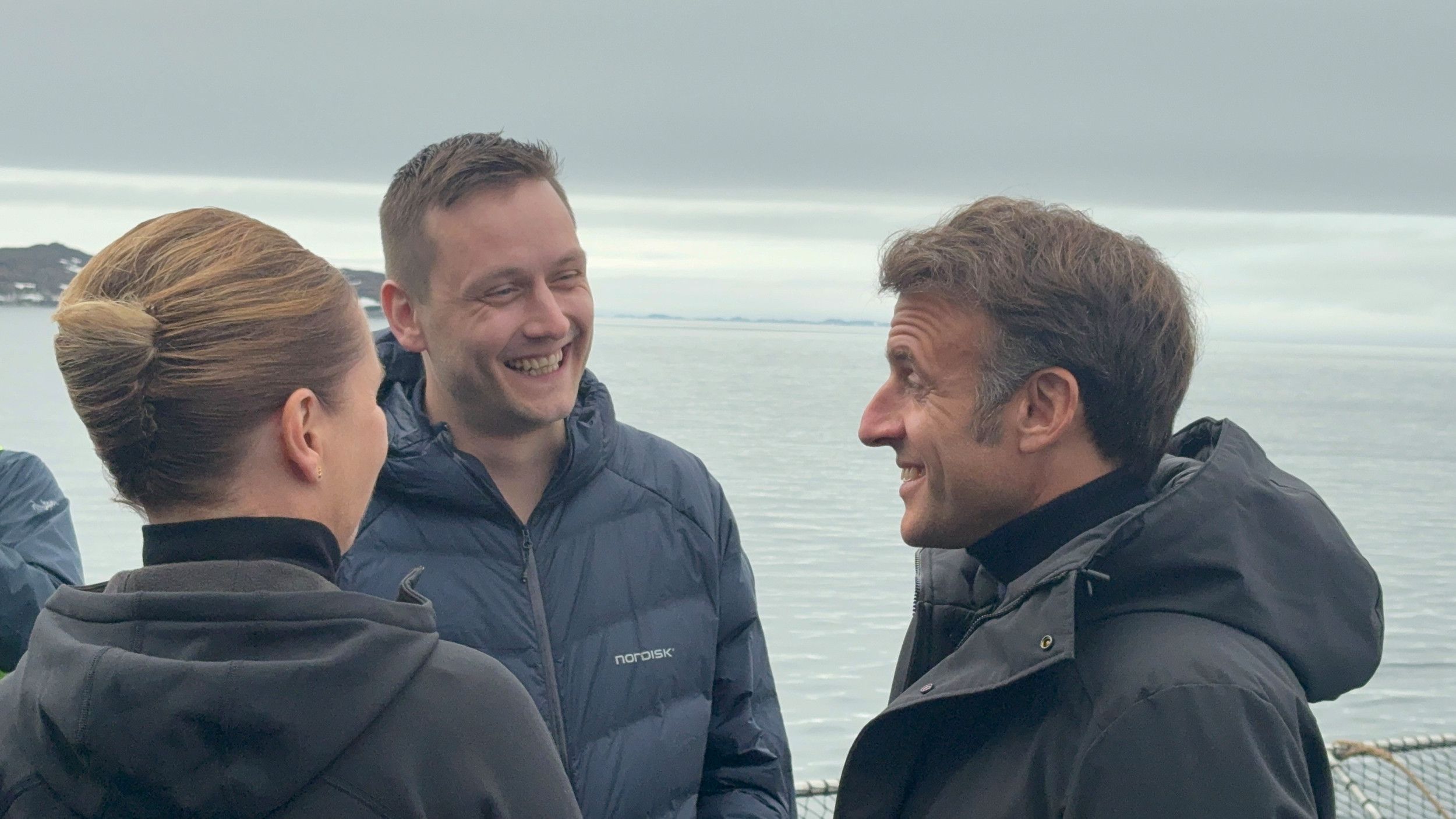 Twee mannen en een vrouw praten lachend met elkaar. Op de achtergrond is zee te zien.