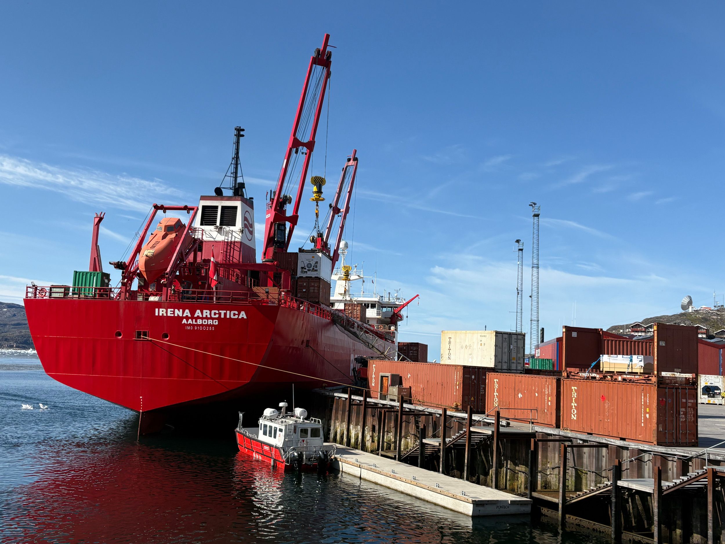 Een rood vrachtschip ligt aan de kade, waar containers wachten om ingeladen te worden.