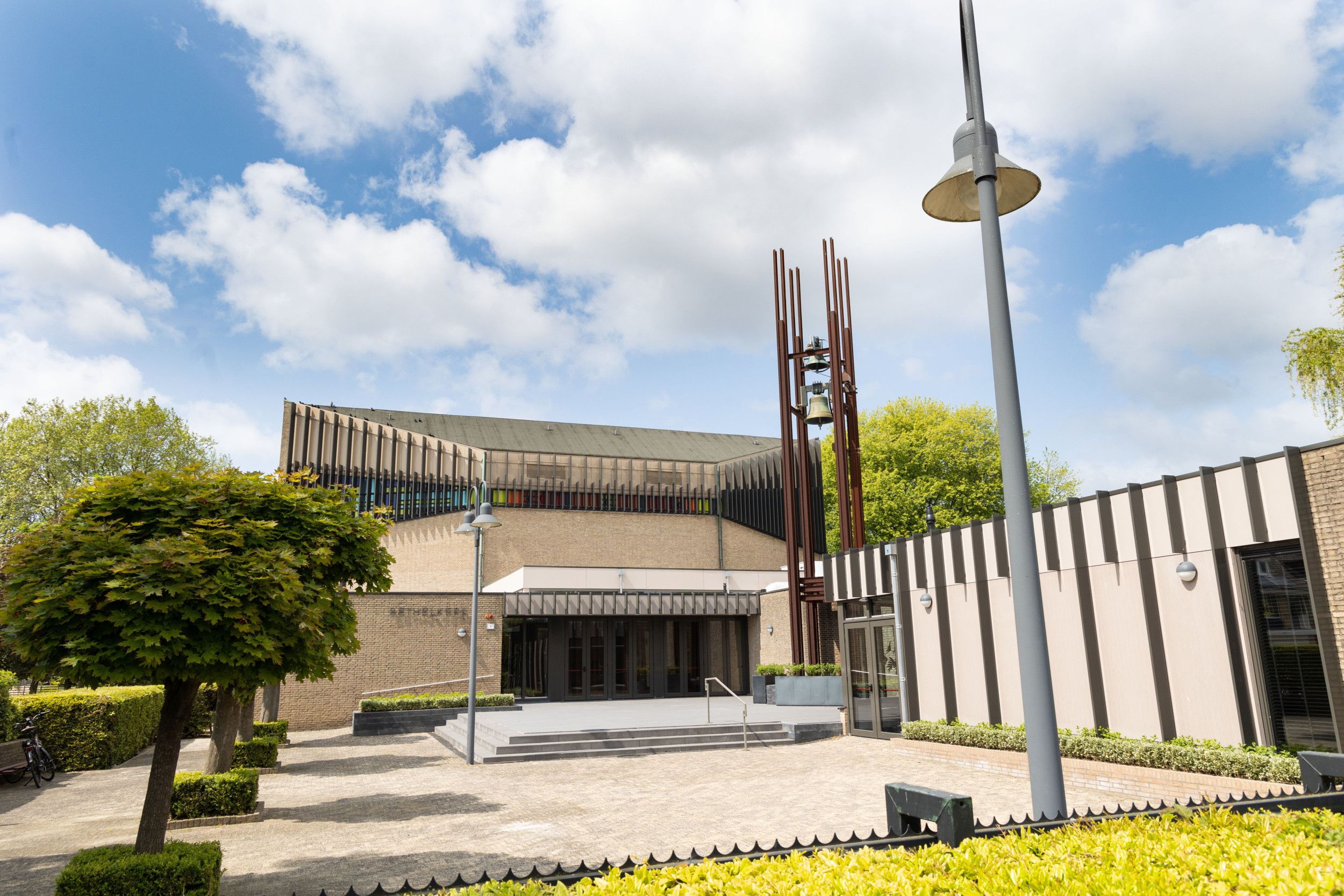 Een modern kerkgebouw, met een stalen klokkentoren. Vooraan zalen.