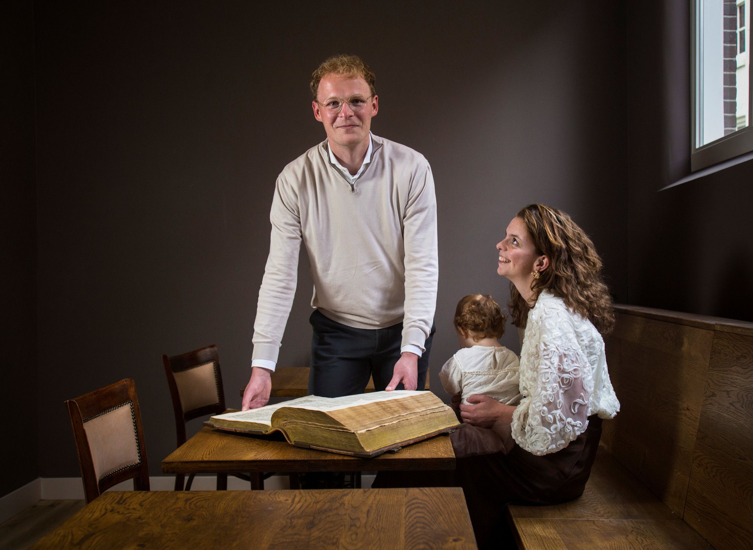 Een jonge man staat achter een tafel met een oude Bijbel, een vrouw met een klein meisje zit ernaast.
