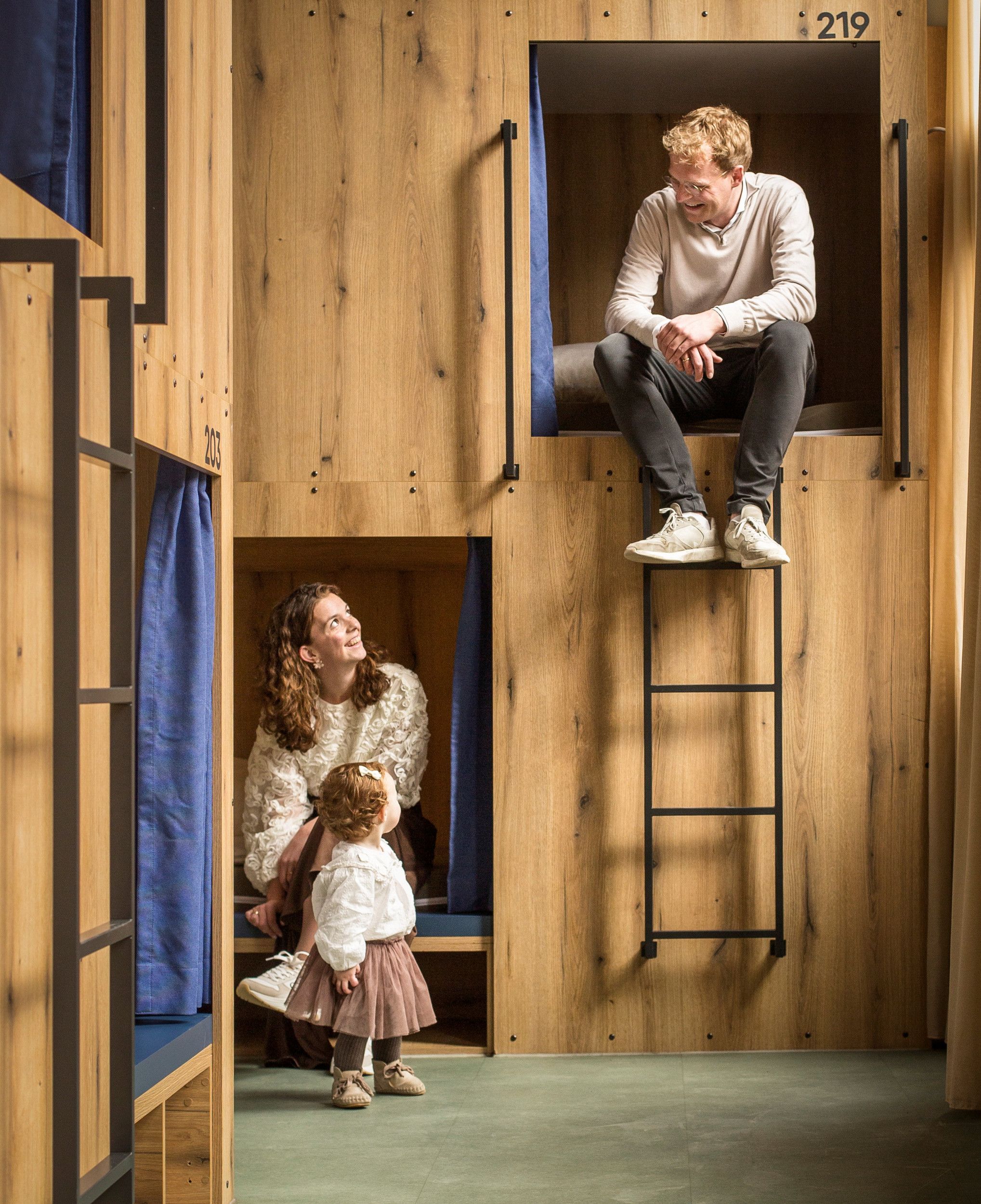 Een jonge vrouw met haar dochtertje zit in een houten slaappod, een jonge man op de trap rechts daarboven.