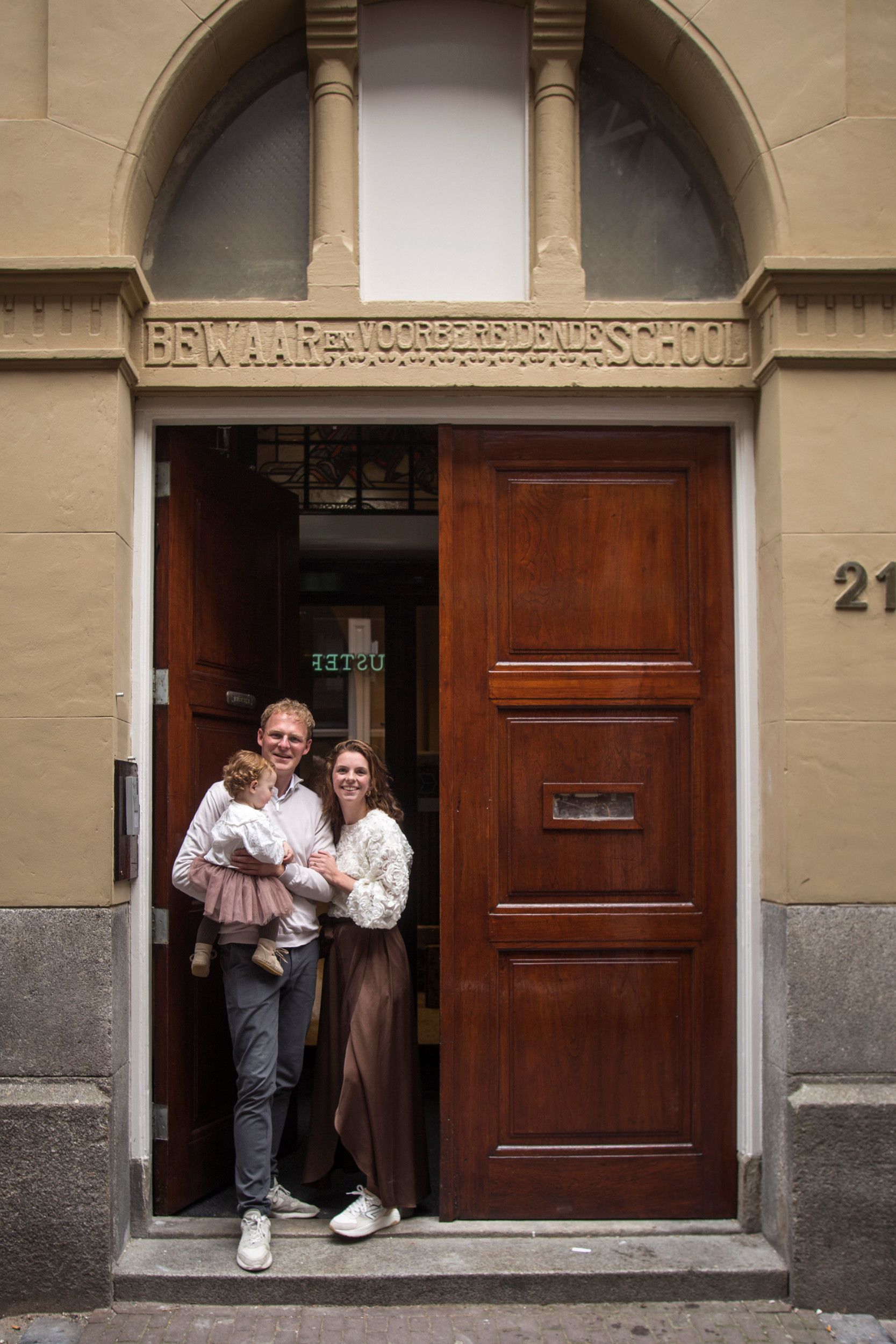 Een jonge vrouw en man, met een klein meisje op de arm, in de deuropening. Daarboven in steen: „Bewaar en voorbereidende school”. 