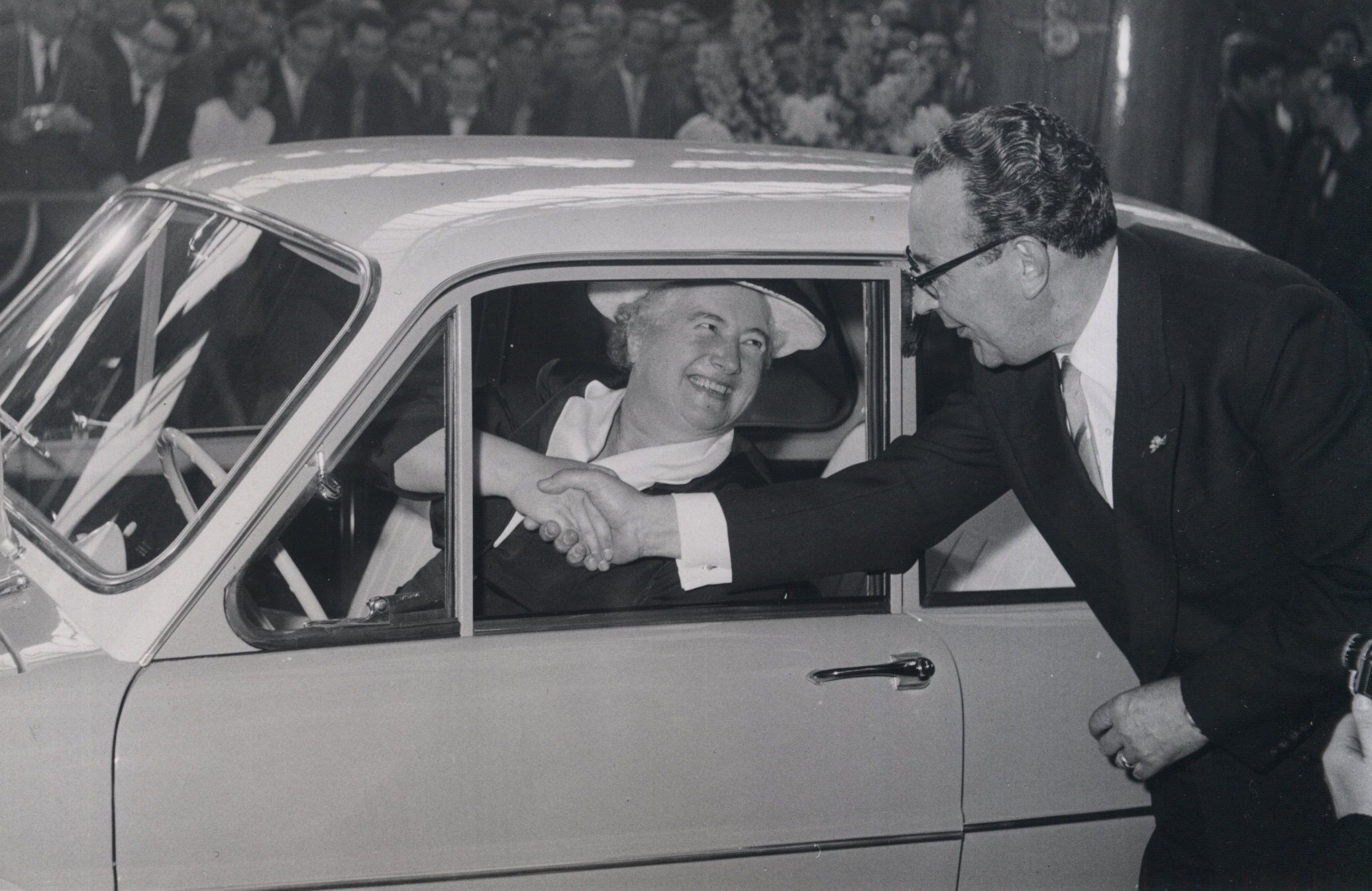 Zwart-wit foto van man in pak met golvend haar die een vrouw met hoed de hand schudt. Ze zit in een kleine auto met het raam open. 