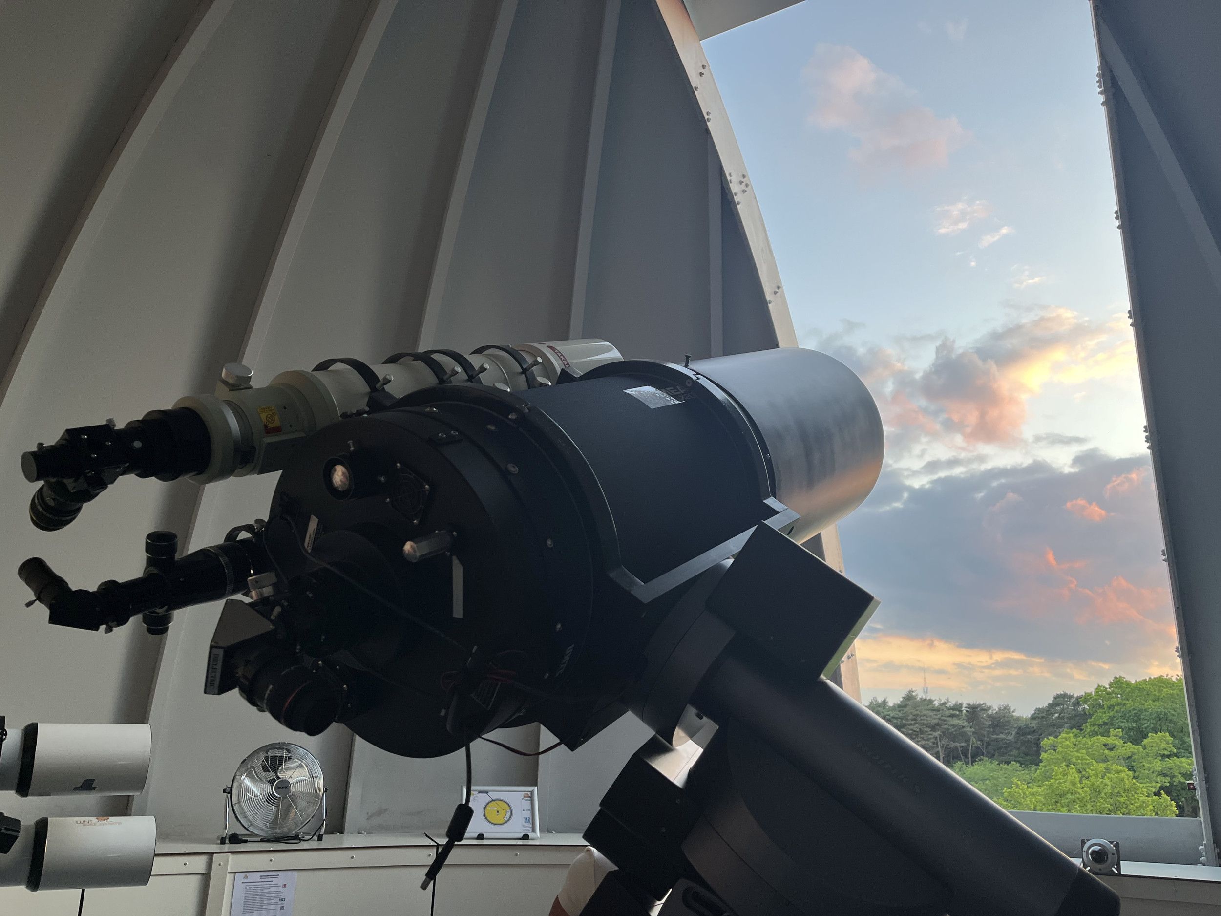 Telescoop in een koepelvormige ruimte gericht op een verticale opening. Buiten zijn bossen te zien met daarboven donkere wolken waartegen de zon schijnt.