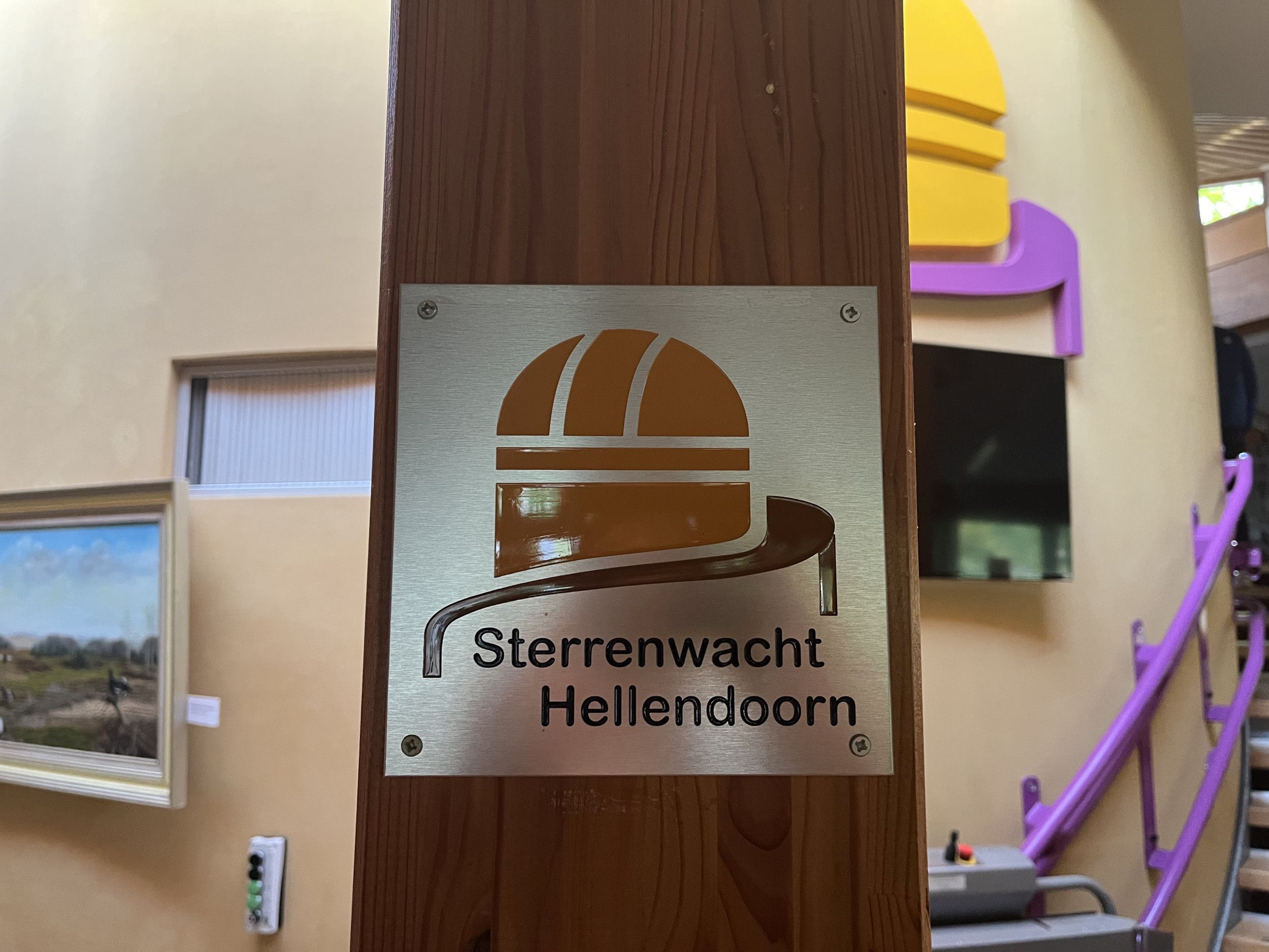 Zilverkleurige plaat met gestileerde koepel en de tekst ”Sterrenwacht Hellendoorn”. De plaat zich vastgeschroefd op een houten plank in een binnenruimte.