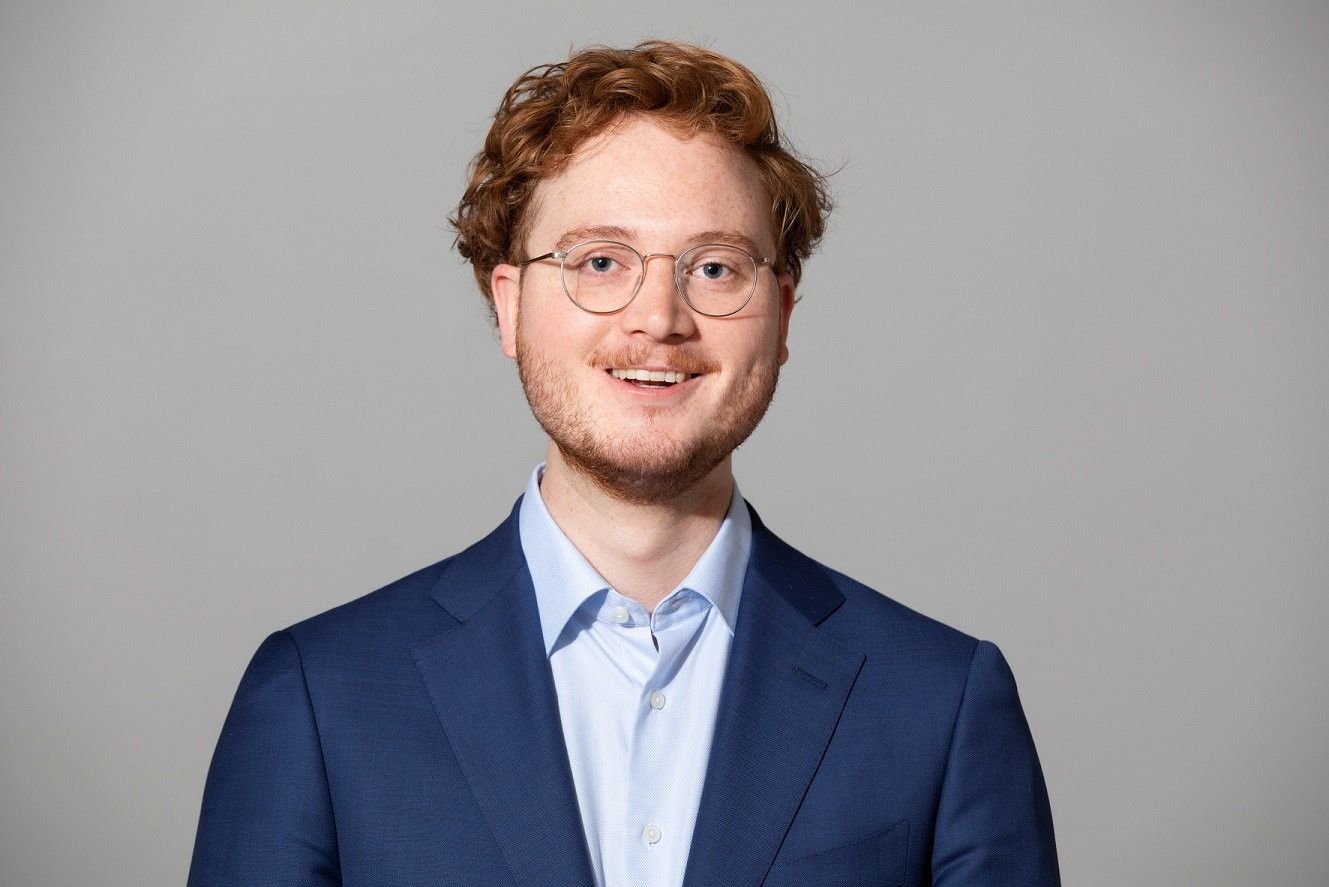 Een jonge man met rossig haar en een bril kijkt in de camera. Hij draagt een blauw colbert met daaronder een lichtblauwe blouse.