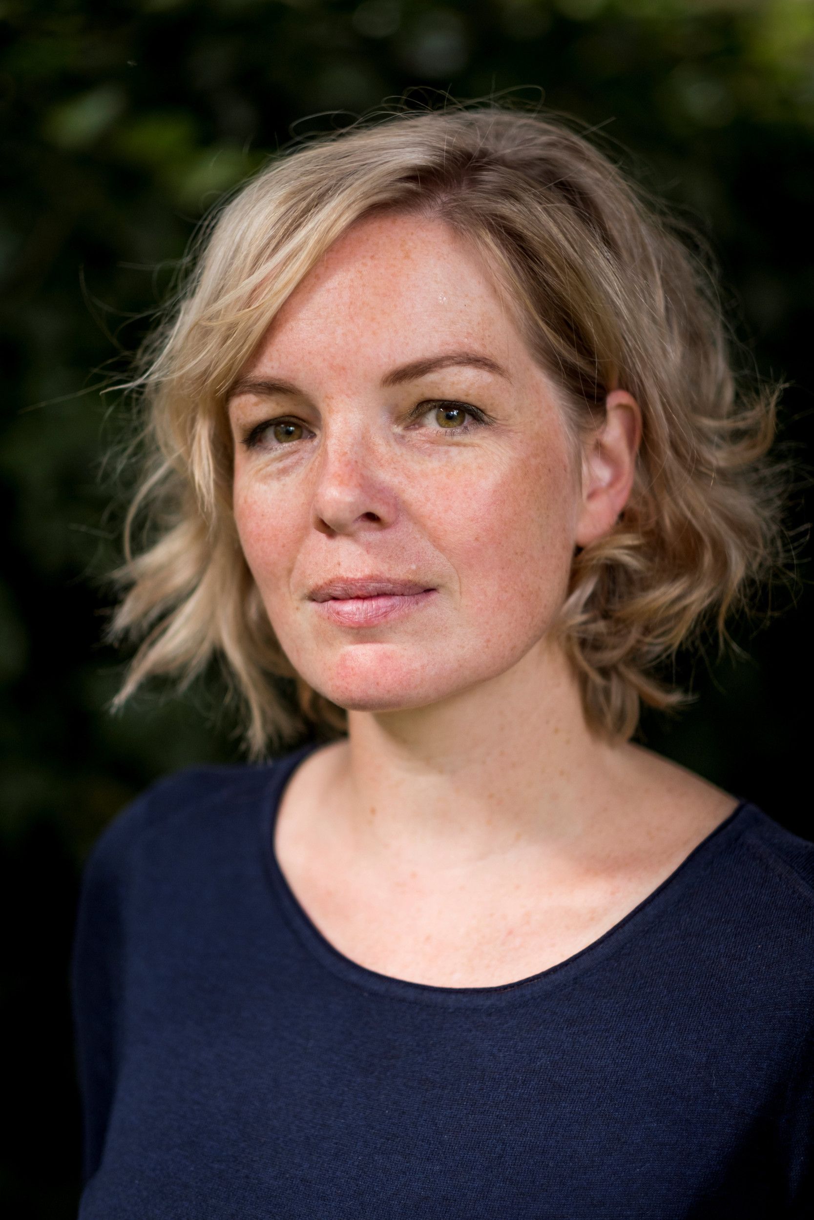 Een vrouw met blond haar en een blauwe trui aan. Ze kijkt in de camera. De achtergrond is zwart.