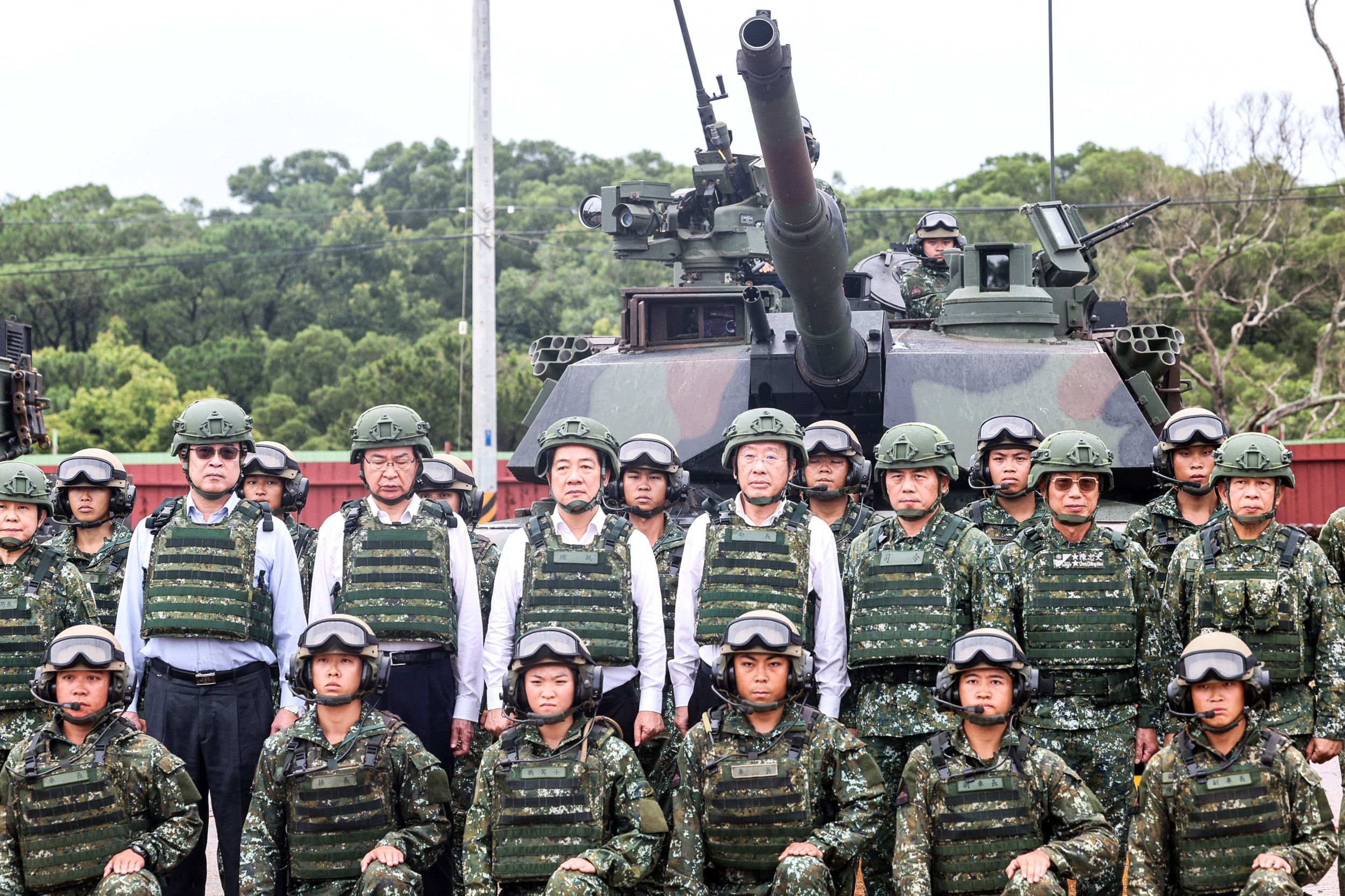 Militairen en politici poseren voor een tank, tegen groene achtergrond.