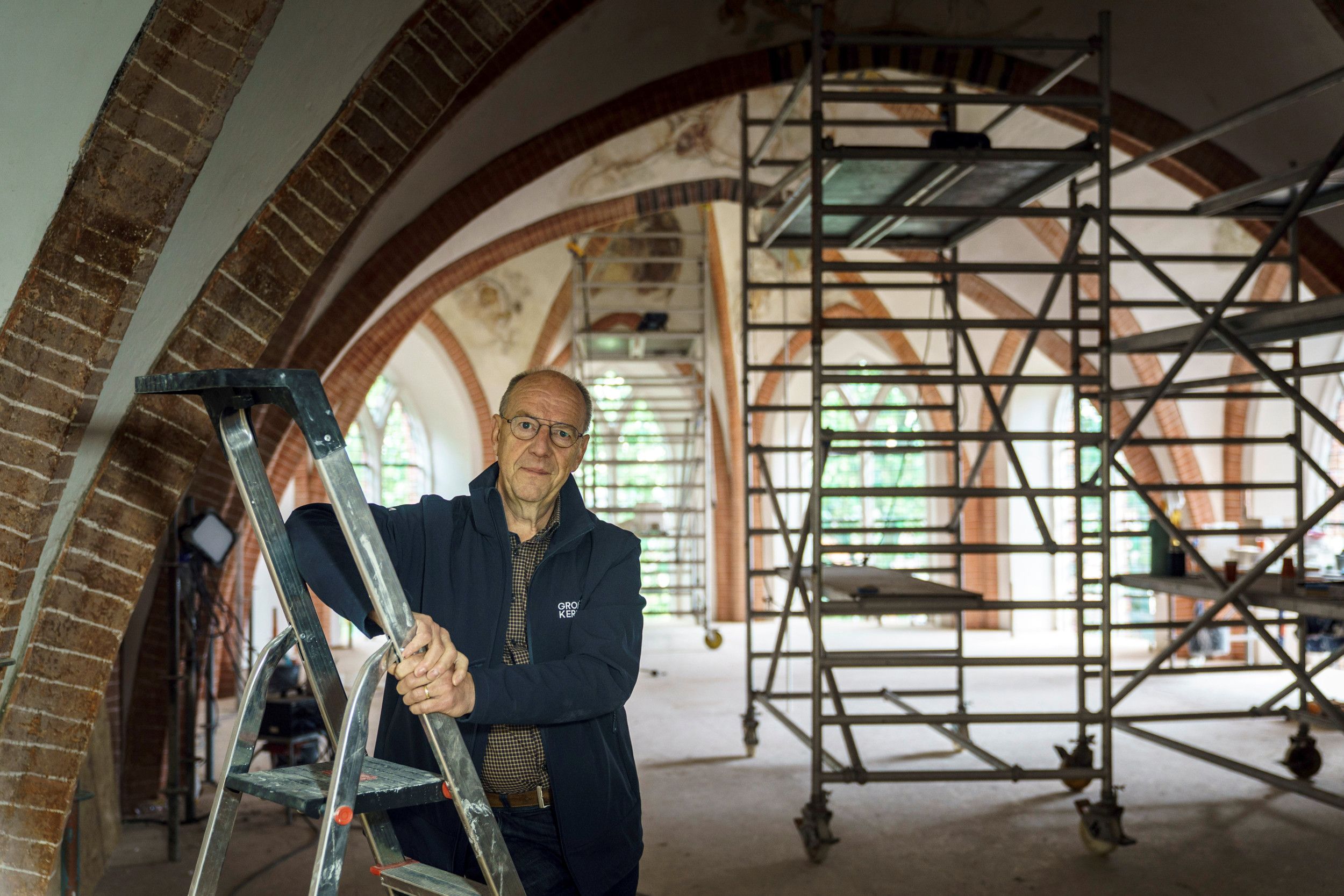 Een oudere man, op een trapje in een kerk. De kerk staat vol steigers.