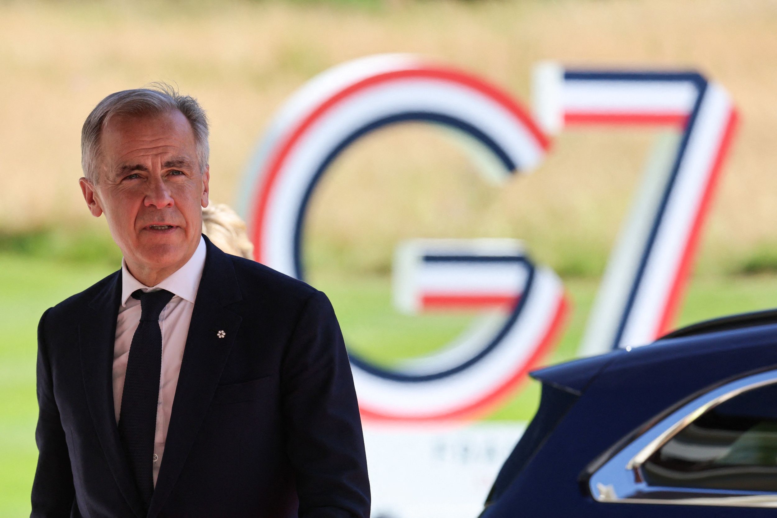 Man met grijs haar, keurig in pak, met op de achtergrond de letters G7 in rood, wit en blauw.