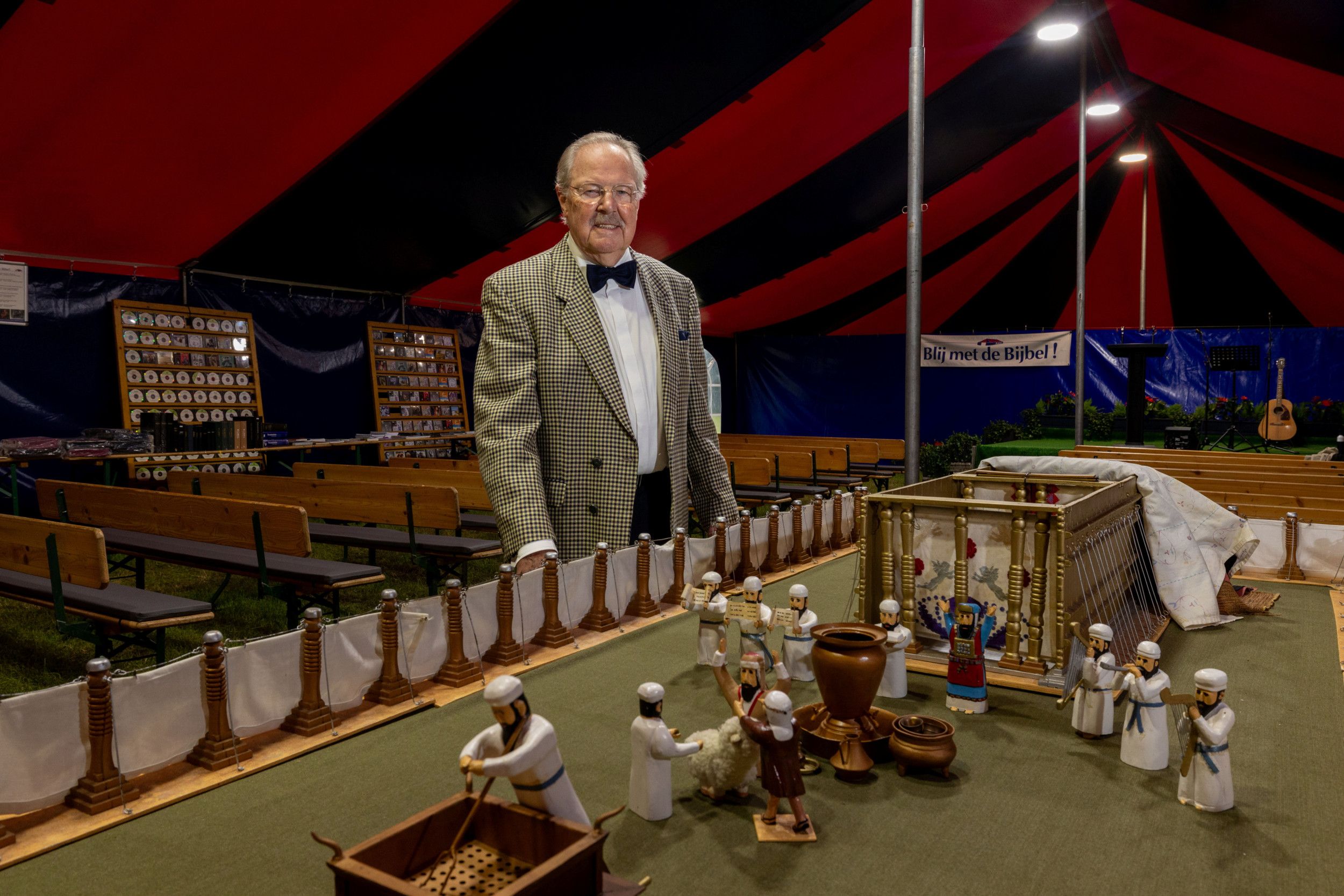 Een man met overhemd, colbert en vlinderstrik staat bij een maquette van de tabernakel in een grote tent, met op de achtergrond houten banken.