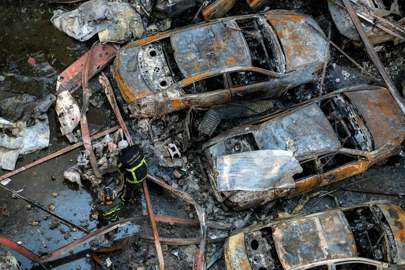 Drie verbrande auto's met daarbij mannen in zwarte pakken met gele strepen, vanboven gefotografeerd.