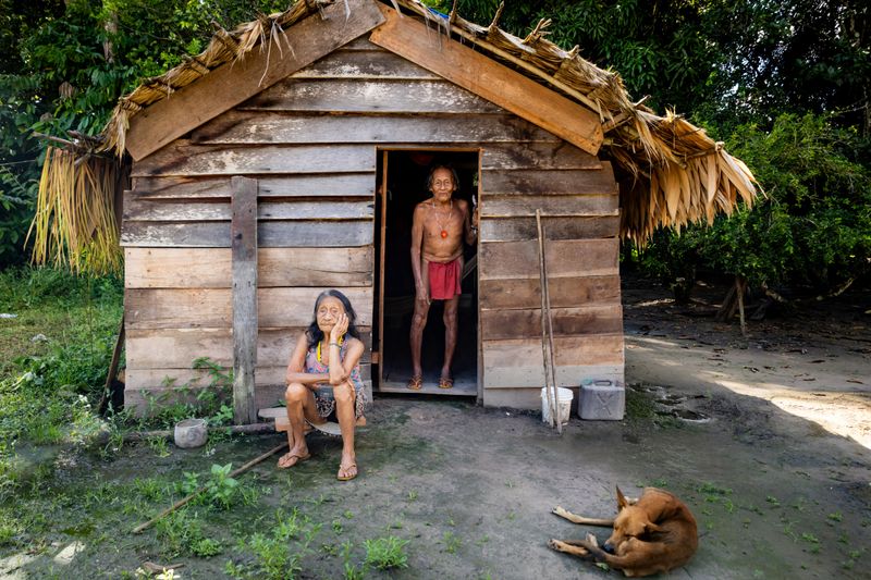Oudere man en vrouw, samen met hun hond, voor hun houten huis met palmbladeren